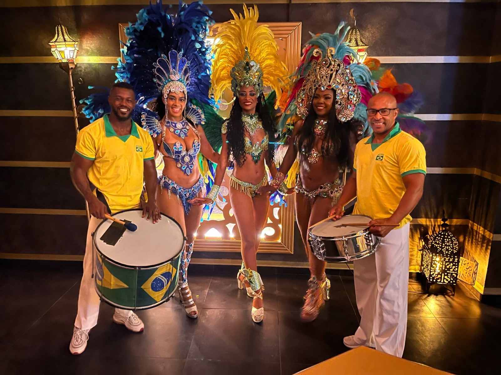 Drei Sambatänzerinnen in Federkostümen zwischen zwei Trommlern in gelben Brasil-Shirts in einem Eventraum
